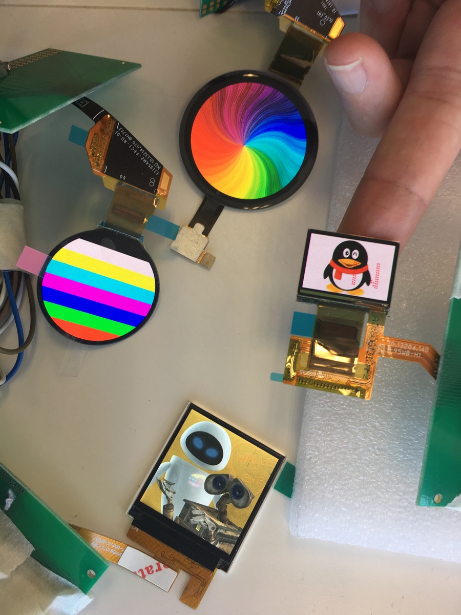 Small round and rectangular colour AMOLED display modules on flexible PCB tails, laid out on a bench showing test patterns