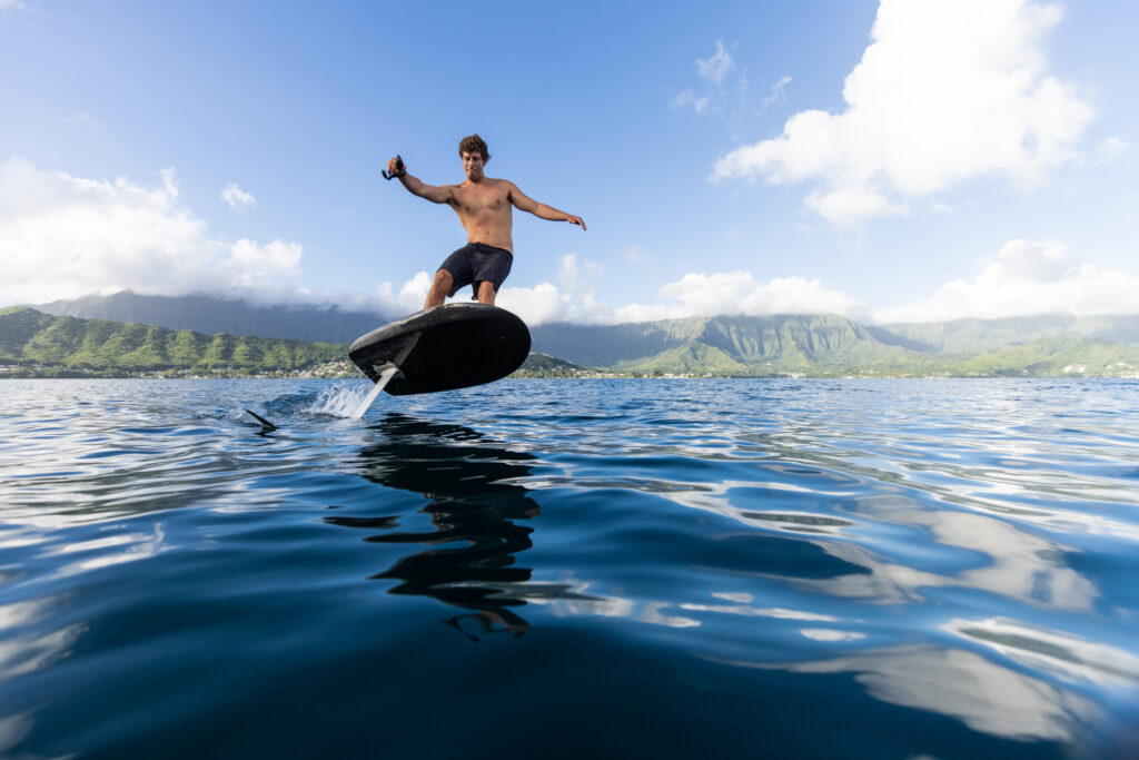 Rider holding the hand controller flies a Fliteboard above calm water, green mountains and cloud in the distance