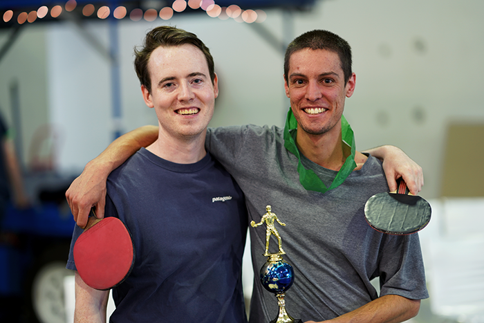 The tournament winner and runner-up arm in arm, holding their table tennis bats and a gold trophy under fairy lights