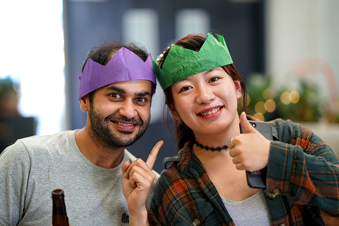 Two colleagues in purple and green paper party crowns, one pointing skyward and the other giving a thumbs up