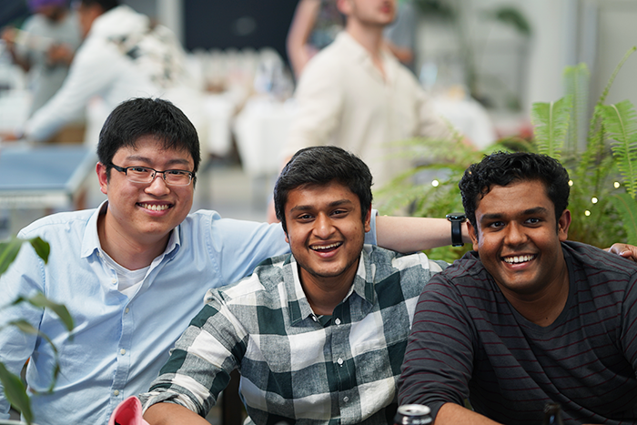 Three colleagues seated together and smiling, with ferns beside them and the end-of-year party carrying on behind