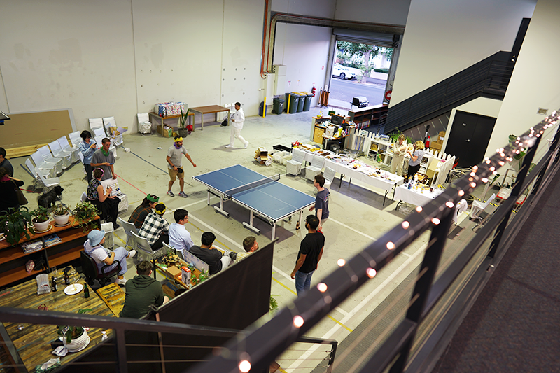 Balcony view of the warehouse office, a table tennis match in play while colleagues watch beside long tables of food