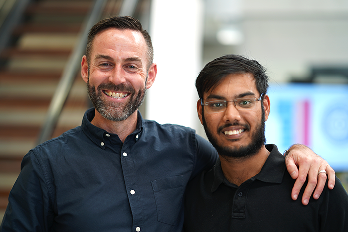 Two colleagues side by side, one with an arm around the other's shoulder, in front of a staircase at the office party