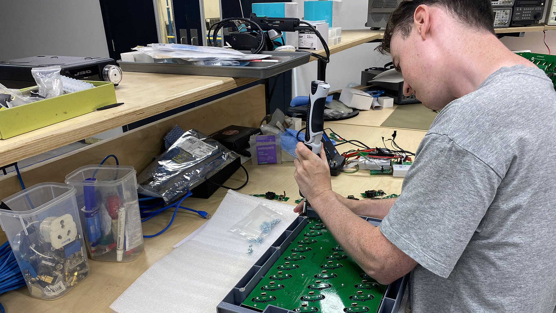Technician at a workbench driving screws into a panel of green circuit boards with an electric screwdriver