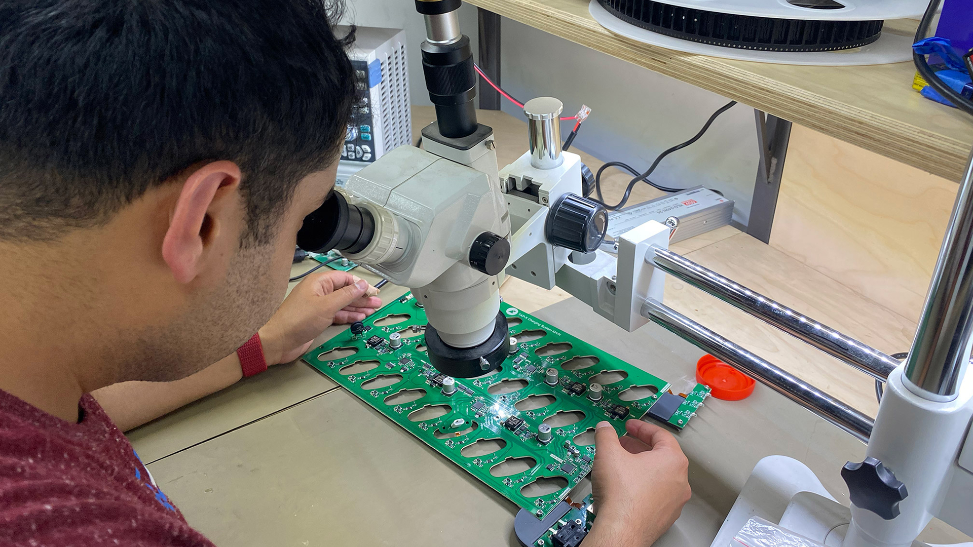 Engineer inspecting a green charging-system circuit board under a stereo microscope at an electronics bench