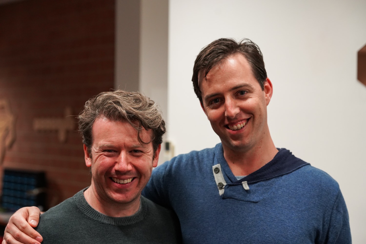 Two smiling guests posing arm in arm at the anniversary night, with a brick wall behind them