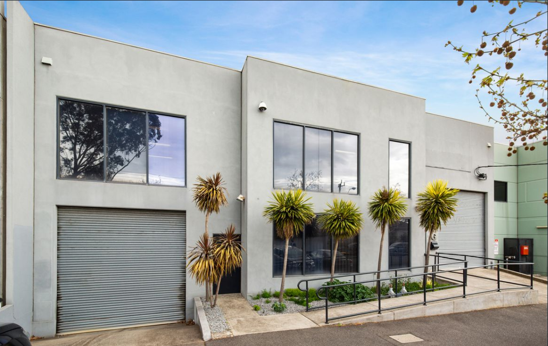 The new Tekt Industries headquarters: a two-storey grey building with roller-door workshop bays, tall windows and cabbage-tree palms lining the ramped entrance