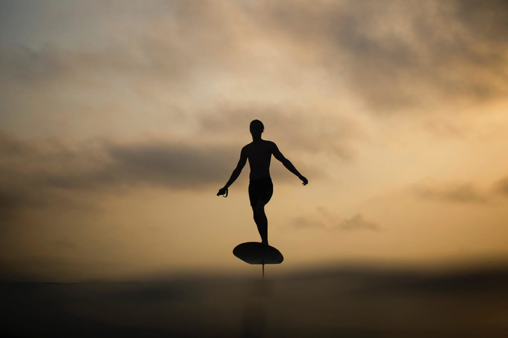 A rider silhouetted against a sunset sky, gliding above the water on a Fliteboard eFoil, handset in hand